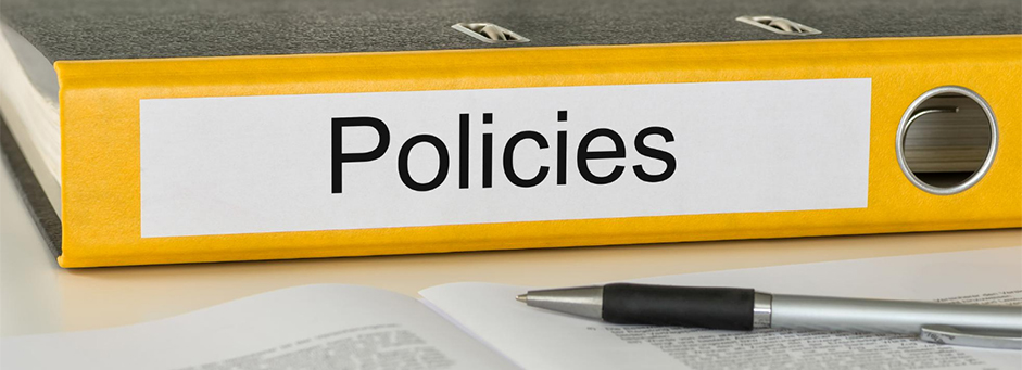 Yellow binder with a white label on the side that says policies with a book laying open in front of the binder with a silver and black pen across it.