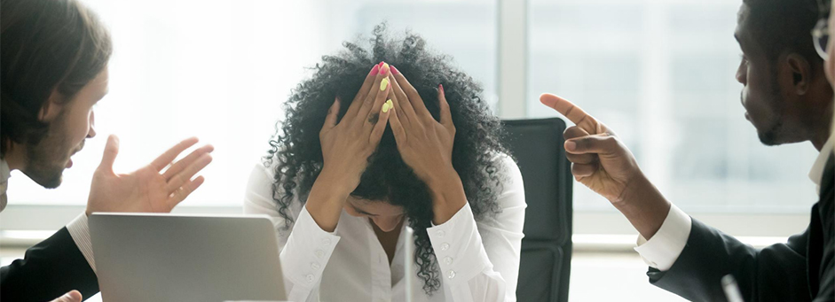 Women with her head down and hands over her head sitting at a table with two mean pointing at her.