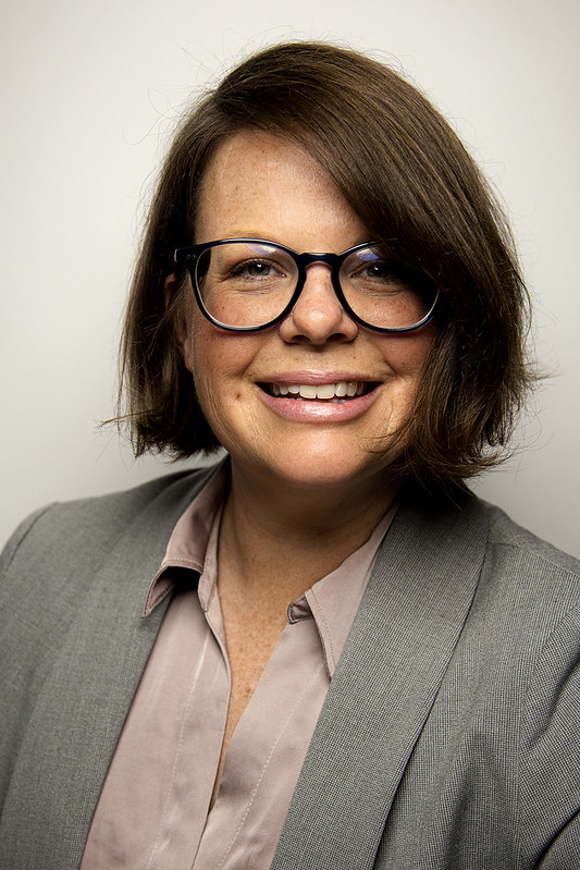 Woman with short dark hair and a gray jacket wearing glasses.